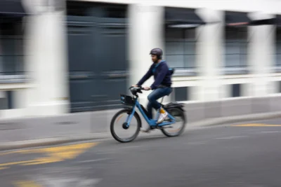 Claude · 5833 x 3889 Cette photographie met en scène un cycliste en mouvement, sujet principal flou suggérant la vitesse. Le cadrage est serré, centré sur le vélo et le cycliste, avec un arrière-plan de colonnes architecturales et une route urbaine.La lumière semble naturelle mais diffuse, peut-être par temps couvert. Les couleurs dominantes sont des tons neutres (gris, bleu) avec des touches de bleu électrique sur le vélo. Le style est dynamique, presque documentaire, capturant l'action avec un effet de vitesse. L'ambiance est urbaine et moderne, évoquant le quotidien actif en ville.