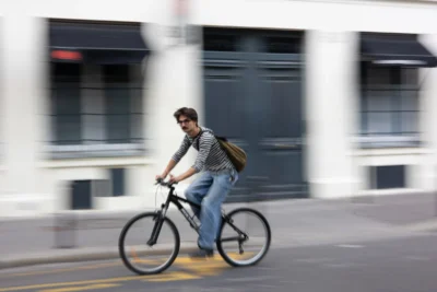 Claude · 5488 x 3659 Cette photographie met en scène un **cycliste** comme sujet principal, en mouvement. La **composition** est dynamique, avec un flou d’arrière-plan suggérant la vitesse. Le **cadrage** est serré, centré sur le cycliste, avec une perspective légèrement basse.La **lumière** semble naturelle, probablement en journée, avec des ombres douces. Les **couleurs dominantes** sont neutres (gris, bleu clair) et contrastées par le bleu du jean et le vert du sac.Le style est **documentaire et spontané**, capturant un instant réel. L’**ambiance** est urbaine et moderne, évoquant la mobilité en ville.