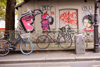 Claude · 5973 x 3982 Cette photographie met en scène des **vélos** attachés à une barrière métallique devant un **mur tagué**. Le sujet principal est l’art urbain coloré, contrastant avec les vélos.La **composition** est équilibrée, avec les vélos en premier plan et les graffitis en arrière-plan. Le **cadrage** est serré, capturant les détails des tags et des bicyclettes.Les **couleurs dominantes** sont vives : rose, bleu, rouge et noir, créant un contraste dynamique. La **lumière** semble naturelle, éclairant les éléments sans ombre marquée.Le **style** est urbain et artistique, évoquant une ambiance **jeune, créative et vivante**, typique des quartiers animés.