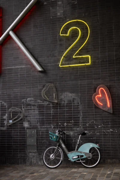 Claude · 3787 x 5681 Cette photographie met en scène un **vélo d’enfant bleu** comme sujet principal, posé devant un mur texturé en briques.La **composition** joue sur les contrastes avec des éléments graphiques : un gros chiffre **2 lumineux jaune**, un cœur néon rouge et des graffitis. Le **cadrage** est serré, centré sur le vélo, avec des éléments urbains en arrière-plan.Les **couleurs dominantes** sont les tons froids (bleu, gris) et les néons vifs (jaune, rouge). La **lumière** est artificielle, créant un effet nocturne et dramatique.Le **style** est urbain et artistique, évoquant une **ambiance mélancolique et poétique**, mêlant nostalgie et modernité.