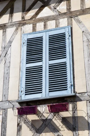 Claude · 3306 x 4959 Cette photographie montre une fenêtre à volets bleus semi-ouverts, style persienne, fixée à une façade de bâtiment en bois et enduit clair. Le sujet principal est cette fenêtre, encadrée par des poutres apparentes en bois sombre.La composition est simple et centrée, avec un cadrage serré mettant en valeur les détails architecturaux. La lumière naturelle éclaire uniformément la scène, créant des ombres douces. Les couleurs dominantes sont le bleu des volets, le blanc crème de l’enduit, et le bois naturel des poutres.L’ambiance est rustique et chaleureuse, évoquant un style traditionnel et campagnard.