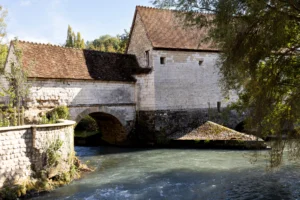 Claude · 5889 x 3926 Cette photographie met en scène un **moulin à eau** en pierre, sujet principal, avec son toit en tuiles et son architecture médiévale. La composition montre un **pont en arc** traversant un ruisseau clair, encadré par une végétation luxuriante.Le cadrage est serré, centré sur le moulin et le cours d’eau. La lumière est naturelle, douce et légèrement latérale, créant des ombres douces. Les **couleurs dominantes** sont les tons beiges et gris de la pierre, contrastant avec le vert des arbres et le bleu-vert de l’eau.Le style est **réaliste et pittoresque**, évoquant une ambiance **pacifique et historique**.