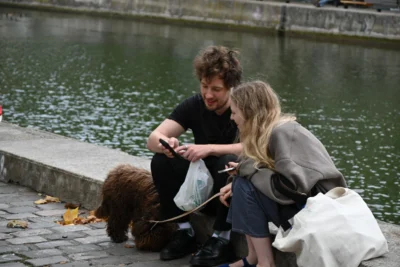 Mary · 13/09/2025 · 5568 x 3712 Cette photographie montre deux personnes assises près d’un plan d’eau, semblant interagir avec un chien. Le sujet principal est cette scène de complicité entre les humains et l’animal.La composition est naturelle, avec un cadrage serré sur les protagonistes et un arrière-plan flou centré sur l’eau. La lumière semble douce et naturelle, probablement en journée. Les couleurs dominantes sont des tons neutres (gris, bleu de l’eau) et quelques touches de couleurs vives comme le jaune des feuilles mortes.Le style est réaliste et spontané, créant une ambiance paisible et chaleureuse.