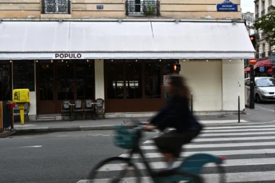 Mary · 13/09/2025 · 2784 x 1856 Cette photographie met en scène un cycliste flou en mouvement devant un restaurant nommé "Populo". La composition joue sur le contraste entre le sujet net (le restaurant) et le cycliste en motion blur. Le cadrage est serré, centré sur la façade du bâtiment.La lumière est naturelle, probablement en journée, avec des ombres douces. Les couleurs dominantes sont neutres : beige, gris et bleu, adoucies par des touches de rouge et de vert. Le style est documentaire et urbain, capturant une scène de vie quotidienne dans une ambiance dynamique et réaliste.
