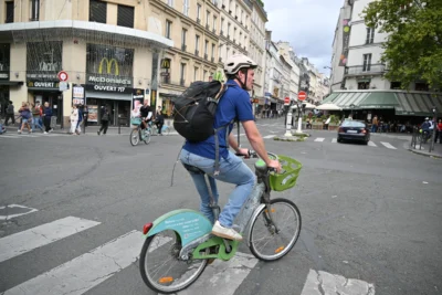 Mary · 13/09/2025 · 5568 x 3712 Cette photographie montre un homme à vélo comme sujet principal, vêtu d'une tenue décontractée avec un casque et un sac à dos. La composition met en avant une scène urbaine animée, avec des piétons et des véhicules en arrière-plan.Le cadrage est serré sur le cycliste, mais inclut aussi des éléments architecturaux et commerciaux (enseignes McDonald's, bâtiments). La lumière semble naturelle, légèrement voilée, créant une ambiance claire mais pas éblouissante. Les couleurs dominantes sont les tons neutres (gris de la route, beige des bâtiments) avec des touches de bleu (vélo) et vert (panier).Le style est documentaire, capturant un moment du quotidien. L'ambiance est dynamique et réaliste, typique d'une ville active.