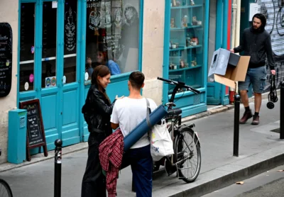Mary · 13/09/2025 · 2784 x 1931 Cette photographie capture une scène urbaine animée. Le sujet principal semble être un groupe de trois personnes près de vélos, dont l’une porte un sac à dos et une veste colorée. La composition montre une rue étroite avec des vitrines colorées en bleu turquoise en arrière-plan.Le cadrage est serré sur les personnages et leurs interactions. La lumière est naturelle, légèrement tamisée, créant des ombres douces. Les couleurs dominantes sont les tons froids (bleu turquoise) et chauds (vêtements rouges et noirs). Le style est réaliste et documentaire, évoquant une ambiance quotidienne et vivante dans une ville européenne.