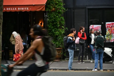 Mary · 13/09/2025 · 2784 x 1856 Cette photographie capture une scène urbaine animée. Le sujet principal est un cycliste en mouvement, flou, suggérant la vitesse. En arrière-plan, des personnes attendent devant un restaurant nommé "A DELLE BIBITE", avec une ambiance détendue.La composition joue sur les contrastes entre le flou dynamique du cycliste et les figures nettes des piétons. Le cadrage est serré, centré sur la rue et les interactions humaines. La lumière est naturelle, probablement en journée, avec des ombres douces.Les couleurs dominantes sont des tons neutres (gris, beige) adoucis par des touches de vert (végétation) et de rouge (enseigne). Le style est documentaire, réaliste, reflétant une ambiance quotidienne et vivante.