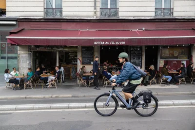 Muriel · 13/09/2025 · 2048 x 1365 Cette photographie met en scène un cycliste comme sujet principal, vêtu d’une veste bleue, roulant sur une piste cyclable en ville.La composition montre une rue urbaine avec des cafés en terrasse occupés, créant un cadrage dynamique. L’arrière-plan inclut des tables et des clients, ajoutant de la profondeur.La lumière est naturelle et douce, typique d’une journée ensoleillée. Les couleurs dominantes sont les tons neutres (gris de la route, bleu du ciel) et les touches chaudes (marron des façades, rouge des enseignes).Le style est réaliste et documentaire, capturant une scène de vie quotidienne. L’ambiance est animée et détendue, reflétant une ville active et conviviale.