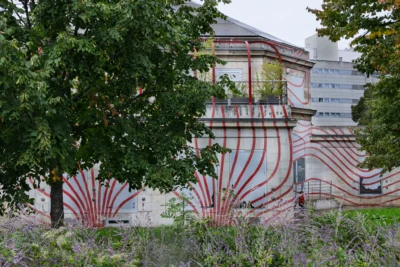 Muriel · 13/09/2025 · 2048 x 1365 Cette photographie met en scène un bâtiment dont la façade est partiellement recouverte d’une installation artistique en forme de vagues ou de rubans rouges. Le sujet principal est cette structure colorée, contrastant avec le gris du mur.La composition joue sur le cadrage serré, encadré par des feuilles d’un arbre et des plantes en bas. La lumière semble naturelle, douce mais diffuse, sans ombres marquées. Les couleurs dominantes sont le rouge vif de l’installation, le gris du bâtiment et les verts des végétaux.Le style est épuré et contemporain, créant une ambiance à la fois artistique et mystérieuse, entre urbanité et nature.