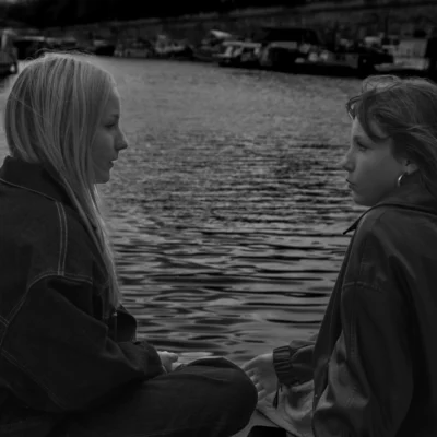 Jean-Paul · 13/09/2025 · 3000 x 3000 Cette photographie en noir et blanc met en scène deux jeunes femmes assises près de l’eau. Le sujet principal est leur interaction, captée en plan serré. La composition est équilibrée, avec un cadrage centré sur leurs visages et épaules.La lumière douce, probablement naturelle, crée des contrastes subtils. Les couleurs dominantes sont les nuances de gris, renforçant une ambiance intime et mélancolique. Le style est sobre et réaliste, évoquant une scène naturelle et authentique. L’ambiance est calme et contemplative, renforcée par le reflet de l’eau et l’arrière-plan discret.