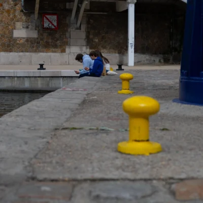 Jean-Paul · 13/09/2025 · 3000 x 3000 Cette photographie montre deux enfants assis près d’un bassin, en arrière-plan. Le sujet principal est l’espace urbain avec ses éléments architecturaux et les bollards jaunes en premier plan.La composition est symétrique avec les bollards alignés, créant une perspective menant vers les enfants. Le cadrage est serré, mettant en valeur les textures des pavés et des murs.Les couleurs dominantes sont les tons gris des pavés et du bassin, contrastés par le jaune vif des bollards. La lumière semble naturelle, douce, et l’ambiance est calme et quotidienne, évoquant un moment de pause dans un environnement urbain.