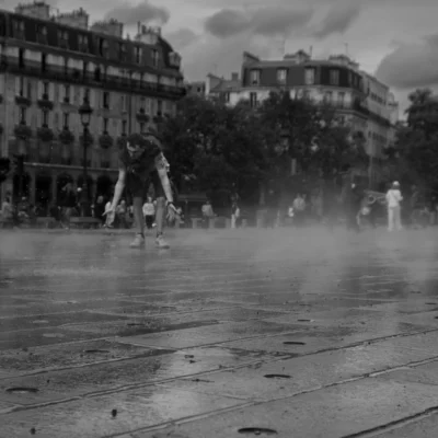 Jean-Paul · 13/09/2025 · 3000 x 3000 Cette photographie en noir et blanc met en scène un enfant comme sujet principal, patinant sur une place mouillée. La composition montre une perspective large avec des bâtiments classiques en arrière-plan et des passants flous.Le cadrage est dynamique, centré sur l'action. La lumière semble naturelle, avec des contrastes doux, accentuant les détails et les mouvements. Les couleurs dominantes, bien que monochromes, jouent sur les nuances de gris et de noir, renforçant l’ambiance dramatique et poétique.Le style est documentaire et spontané, évoquant une scène de vie urbaine. L’ambiance est à la fois joyeuse et mélancolique, reflétant la fraîcheur d’un moment éphémère.