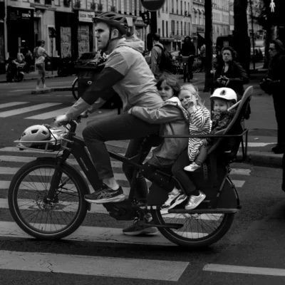 Jean-Paul · 13/09/2025 · 3000 x 3000 Cette photographie en noir et blanc met en scène un homme transportant deux enfants dans un siège arrière fixé à un vélo. Le sujet principal est cette famille en mouvement, capturée en pleine ville.La composition est dynamique, avec un cadrage serré sur le vélo et ses occupants, tandis que l’arrière-plan flou montre des passants et des bâtiments. La lumière semble naturelle, probablement en journée, avec des contrastes marqués. Les tons dominants sont les gris variés, typiques du noir et blanc, renforçant l’ambiance urbaine et intemporelle. Le style est documentaire, authentique et spontané, évoquant une scène du quotidien.