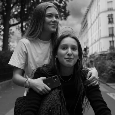 Jean-Paul · 13/09/2025 · 3000 x 3000 Cette photographie met en scène deux jeunes femmes en extérieur. Le sujet principal est leur complicité, avec la personne devant tenant un smartphone et l’autre souriante derrière elle, les bras autour de ses épaules.La composition est naturelle et serrée, avec un cadrage rapproché. La lumière est douce, légèrement tamisée, créant des contrastes mous. Les couleurs dominantes sont les tons de gris et noir, typiques d’un rendu monochrome.Le style est documentaire et spontané, évoquant une ambiance intime et chaleureuse, presque nostalgique.