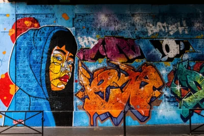 Pascal N. · 13/09/2025 · 3240 x 2160 Cette photographie montre un **mural urbain** comme sujet principal. La composition met en avant un **visage stylisé** à gauche, partiellement caché par une capuche, entouré de motifs colorés et abstraits.Le cadrage est serré, capturant les détails des graffitis. La lumière naturelle éclaire le mur, créant des contrastes. Les **couleurs dominantes** sont des **tons vifs** (bleu, orange, violet) et des dégradés.Le style est **graffiti artistique**, mêlant réalisme et abstraction. L’ambiance est **urbaine et dynamique**, évoquant la culture street art et une touche de mystère.