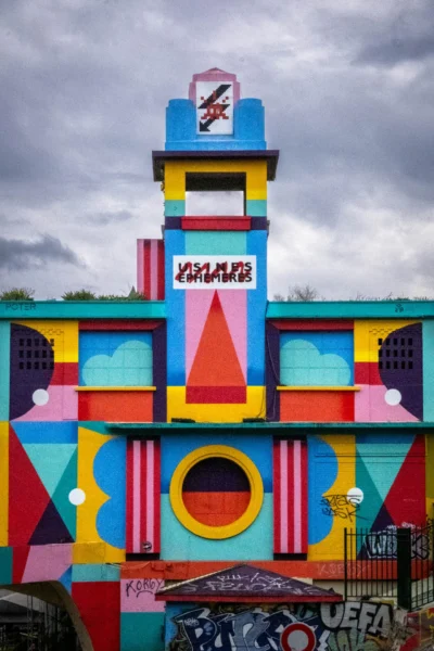 Pascal N. · 13/09/2025 · 1440 x 2160 Cette photographie montre une structure colorée et géométrique, évoquant un bâtiment ou une installation artistique. Le sujet principal est ce mur aux formes anguleuses et aux couleurs vives, dominées par des tons de bleu, jaune, rouge et vert.La composition est dynamique, avec des éléments superposés et des formes variées. Le cadrage est serré, mettant en valeur les détails architecturaux. La lumière semble naturelle, avec des contrastes doux. L’ambiance est festive et ludique, rappelant un style pop art ou street art. Les nuages dans le ciel ajoutent une touche dramatique.