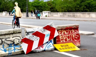 Pascale · 13/09/2025 · 2500 x 1482 Cette photographie montre des panneaux de signalisation renversés et tagués, avec des inscriptions comme "ÉPOTA" et "RUE BARRIÈRE". Le sujet principal est ces panneaux détériorés, placés sur une chaussée.La composition met en avant un avant-plan désordonné, contrastant avec l’arrière-plan où l’on voit des passants et une route. Le cadrage est serré sur les panneaux, avec une légère profondeur de champ.Les couleurs dominantes sont les tons gris de la route, les teintes vives des tags (rouge, bleu) et le jaune du panneau "RUE BARRIÈRE". La lumière semble naturelle, avec des ombres douces. L’ambiance est urbaine et chaotique, évoquant un acte de vandalisme ou de protestation.