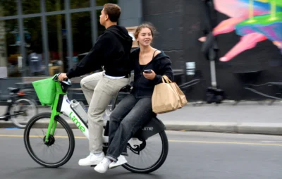 Pascale · 13/09/2025 · 2500 x 1584 Cette photographie montre un couple en mouvement sur un vélo électrique en ville. Le sujet principal est la femme assise, tenant un sac, tandis que l’homme pédale derrière elle.La composition est dynamique, avec un cadrage serré sur les deux personnages. La lumière semble naturelle, bien que légèrement contrastée, mettant en valeur les contours. Les couleurs dominantes sont les tons neutres (gris, noir) et quelques touches vives comme le vert du panier et le bleu du sac.Le style est documentaire et spontané, capturant un moment du quotidien. L’ambiance est urbaine et moderne, évoquant une vie active et connectée.