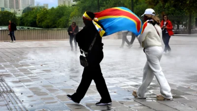 Pascale · 13/09/2025 · 2500 x 1413 Cette photographie capture deux personnes marchant sur une place pavée. Le sujet principal est un homme en costume noir tenant un parapluie coloré (arc-en-ciel), tandis qu’une femme en tenue blanche le suit. La composition est dynamique, avec un cadrage serré sur les deux personnages en mouvement.La lumière semble naturelle, légèrement diffuse, créant des contrastes doux. Les couleurs dominantes sont les tons vifs de l'arc-en-ciel et les contrastes entre le noir du costume et le blanc de la tenue. Le style est documentaire et spontané, évoquant une ambiance urbaine et moderne, presque poétique malgré le contexte quotidien.