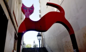Pascale · 06/09/2025 · 700 x 424 Cette photographie met en scène une sculpture moderne représentant une tête de chat stylisée, aux couleurs vives. Le sujet principal est ce chat rouge et bleu, suspendu au-dessus d’un escalier en pierre.La composition joue sur le contraste entre l’art contemporain et l’architecture ancienne. Le cadrage est serré, mettant en valeur la sculpture tout en incluant partiellement l’escalier et un lampadaire. La lumière, naturelle et douce, accentue les teintes saturées. Les couleurs dominantes sont le rouge, le bleu et le blanc, créant une ambiance à la fois dynamique et mystérieuse, mêlant modernité et tradition.