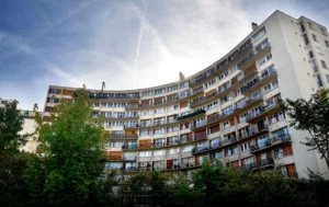 Pascale · 06/09/2025 · 700 x 442 Cette photographie montre un immeuble résidentiel moderne aux lignes courbes, sujet principal. La composition met en valeur sa façade avec des balcons en saillie et des fenêtres alignées. Le cadrage, légèrement bas, accentue la hauteur de l'édifice.La lumière, naturelle et douce, semble provenir d’un ciel partiellement nuageux. Les couleurs dominantes sont les tons beiges, blancs et bruns, adoucis par des touches de vert des arbres en bas. Le style architectural est fonctionnel et épuré, évoquant une ambiance urbaine paisible et contemporaine.