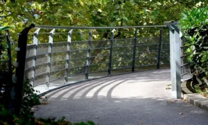 Pascale · 06/09/2025 · 700 x 419 Cette photographie montre un **pont métallique enjambant un chemin de terre**. Le **sujet principal** est la structure du pont, avec ses **barrières en métal gris** et ses **poteaux blancs**.La **composition** est symétrique, avec un cadrage serré sur le pont et une légère perspective convergente. L’**ambiance** est calme et naturelle, renforcée par les **feuillages verts** en arrière-plan.La **lumière** semble douce et naturelle, probablement en journée. Les **couleurs dominantes** sont le **gris métallique**, le **vert des arbres** et le **beige de la terre**. Le style est **réaliste et épuré**, évoquant la sérénité d’un espace naturel.