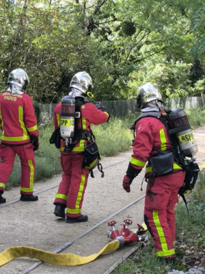 Laurence · 06/09/2025 · 3060 x 4080 Cette photographie montre trois pompiers en tenue de protection, équipés de bouteilles d'air et de casques. Ils semblent en situation d'entraînement ou d'intervention.La composition est dynamique, avec un cadrage serré sur les individus. Les couleurs dominantes sont le rouge vif des combinaisons, le jaune des tuyaux et le vert des arbres en arrière-plan. La lumière naturelle éclaire la scène, créant des ombres douces.Le style est documentaire, capturant une activité professionnelle. L'ambiance est sérieuse et opérationnelle, reflétant la préparation et la vigilance des secours.