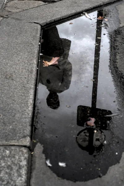 Ramón · 13/09/2025 · 1470 x 2200 Cette photographie montre une personne vue de dessus à travers une flaque d’eau, reflétant son image inversée. Le sujet principal est le pied et la jambe de la personne, ainsi que son reflet déformé.La composition repose sur un cadrage serré et symétrique, centré sur la flaque. La lumière semble naturelle, avec des contrastes doux, créant des reflets sombres et des nuances claires.Les couleurs dominantes sont les tons gris et noirs, avec des touches de blanc et de bleu dans l’eau. Le style est réaliste et documentaire, évoquant une ambiance urbaine et mélancolique, presque poétique.