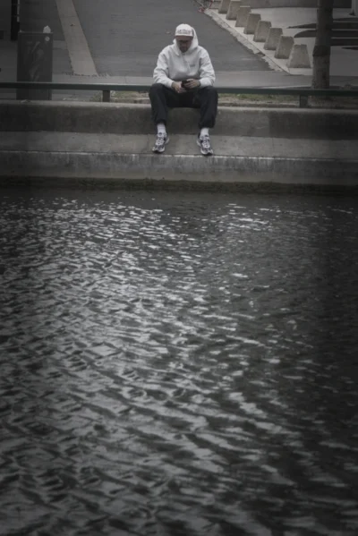 Ramón · 13/09/2025 · 1470 x 2200 Cette photographie met en scène un skateboarder assis sur une margelle, les pieds dans l’eau. Le sujet principal est le jeune homme, vêtu d’un sweat et d’un bonnet, en position détendue.La composition est épurée, avec un cadrage serré sur le sujet et un premier plan dominé par l’eau. La lumière, douce et tamisée, crée des contrastes subtils. Les couleurs, en noir et blanc, renforcent l’ambiance mélancolique et introspective. Le style est documentaire et sobre, évoquant une atmosphère calme et contemplative.