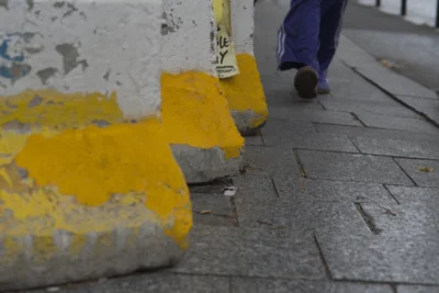 Ramón · 13/09/2025 · 3291 x 2200 Cette photographie met en scène des **barrières de protection jaune et grise** posées sur un trottoir en pavés. Le **sujet principal** semble être ces barrières, partiellement endommagées.La **composition** est simple, avec un **cadrage serré** sur les obstacles, laissant peu de place à d'autres éléments. Un **pied en mouvement** (vêtu de baskets) apparaît en haut à droite, suggérant une personne marchant.Les **couleurs dominantes** sont le **jaune vif** des barrières et le **gris** des pavés et de la peinture écaillée. La **lumière** est naturelle, sans ombre marquée, créant une ambiance **neutre et urbaine**. Le style est **documentaire et réaliste**.