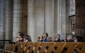 Muriel · 1080 x 677 Cette photographie montre un groupe de personnes assis dans une église, écoutant ou participant à une cérémonie religieuse. Le sujet principal est l’assemblée, vue de dos ou de trois quarts.La composition est horizontale, avec un cadrage serré sur les fidèles et un fond architectural imposant (piliers, vitraux). La lumière, tamisée et douce, provient probablement des vitraux et des luminaires, créant une ambiance solennelle et contemplative. Les couleurs dominantes sont les tons neutres (beige, gris) avec des touches de bleu et rouge dans les vitraux.Le style est réaliste et sobre, évoquant une atmosphère de recueillement et de spiritualité.