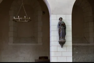 Muriel · 1080 x 729 Cette photographie met en scène une statue religieuse en pierre, probablement une Vierge à l'Enfant, fixée sur un mur en pierre claire. La composition est épurée, avec un cadrage serré centré sur la statue, encadrée par des arches.La lumière est douce et naturelle, créant des ombres subtiles. Les couleurs dominantes sont les tons gris-beige des pierres et les teintes bleutées de la statue. Le style évoque l’architecture classique ou médiévale, avec une ambiance sobre et solennelle, presque sacrée. L’éclairage tamisé accentue la sérénité et la contemplation.