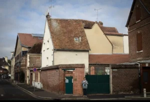 Muriel · 1080 x 731 Cette photographie montre une rue résidentielle avec des maisons en rangée comme sujet principal. La composition met en avant des bâtiments aux façades anciennes, avec des matériaux variés : briques, enduit clair et toits en tuiles.Le cadrage est serré, centré sur les maisons, avec une perspective légèrement inclinée. La lumière est naturelle, douce et diffuse, typique d’un jour nuageux. Les couleurs dominantes sont les tons beiges, bruns et verts, renforçant un style traditionnel et sobre.L’ambiance est calme et rétro, évoquant un quartier historique et paisible.