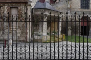 Muriel · 1080 x 720 Cette photographie montre une entrée monumentale, probablement d’un édifice historique ou religieux. Le sujet principal est une porte en pierre ornées de motifs sculptés, entourée d’une grille en fer forgé décorative.La composition met en valeur les détails architecturaux et la texture des matériaux. Le cadrage serré accentue les éléments décoratifs et la profondeur de l’ombre. La lumière, tamisée, crée des contrastes entre les zones éclairées et ombragées, renforçant l’ambiance mystérieuse et solennelle. Les couleurs dominantes sont les tons gris-beige et verts, typiques des pierres anciennes et du métal oxydé. Le style évoque une atmosphère historique et majestueuse.