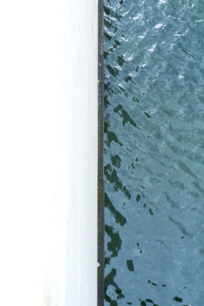 Renato · 13/09/2025 · 2560 x 3840 Cette photographie capture une portion de surface aquatique reflétant le ciel. Le **sujet principal** est l’eau, avec ses reflets bleutés et ses ondulations. La **composition** est minimaliste, avec un **cadrage serré** sur une bande verticale, séparant deux espaces visuels distincts.La **lumière** semble naturelle, douce et diffuse, créant des jeux de transparence. Les **couleurs dominantes** sont des teintes de bleu et de blanc, évoquant une ambiance calme et apaisante. Le **style** est épuré, presque abstrait, tandis que l’**ambiance** est contemplative et sereine, comme une invitation à observer la beauté simple des éléments naturels.