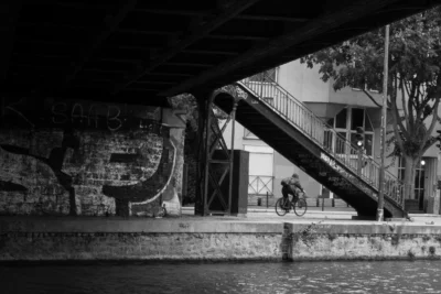 Renato · 13/09/2025 · 2848 x 1899 Cette photographie en noir et blanc met en scène un cycliste comme sujet principal, en mouvement sur un pont. La composition joue sur les lignes architecturales, avec une rampe en béton et un auvent qui encadrent la scène. Le cadrage est serré, centré sur le cycliste et les éléments urbains environnants.La lumière, tamisée, crée des contrastes marqués et accentue les ombres, renforçant l'ambiance contemplative. Les couleurs dominantes, bien que monochromes, exploitent les nuances de gris pour donner du relief. Le style est documentaire et artistique, évoquant une atmosphère mélancolique et urbaine.