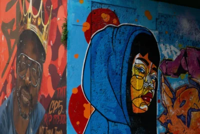 Renato · 13/09/2025 · 3840 x 2560 Cette photographie montre un mural urbain avec deux portraits stylisés comme sujets principaux. À gauche, un homme barbu porte une couronne, tandis qu’à droite, une silhouette féminine aux traits marqués est vêtue d’une cape bleue.La composition est dynamique, avec un cadrage serré sur les visages. Les couleurs dominantes sont vives : bleus, rouges, oranges et jaunes, créant un contraste saisissant. Le style est graffiti, aux traits expressifs et aux aplats colorés.L’ambiance est à la fois puissante et artistique, reflétant une énergie urbaine et culturelle. La lumière semble naturelle, mettant en valeur les détails des visages.
