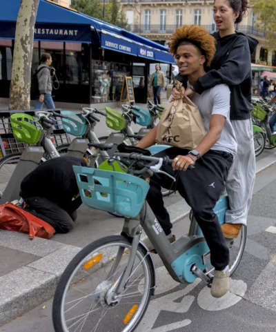 Thibault · 13/09/2025 · 3191 x 3839 Cette photographie met en scène une femme comme sujet principal, assise sur un vélo en libre-service avec panier, en ville. La composition montre une rue urbaine avec des vélos alignés et des passants en arrière-plan.Le cadrage est serré sur la femme, avec une profondeur de champ qui floute légèrement l’arrière-plan. La lumière est naturelle, probablement en journée, avec des ombres douces. Les couleurs dominantes sont les tons neutres (gris, beige) et les touches vives comme le bleu du vélo et le vert des arbres.Le style est documentaire et spontané, capturant une scène de vie quotidienne. L’ambiance est dynamique et urbaine.
