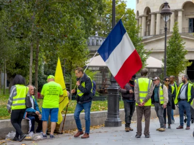 Thibault · 13/09/2025 · 3840 x 2880 Cette photographie montre un groupe de manifestants vêtus de vestes jaunes, emblématiques des "Gilets jaunes". Le sujet principal est un individu tenant le drapeau français. La composition met en scène une scène urbaine avec des arbres et un bâtiment en arrière-plan. Le cadrage est serré sur les manifestants, capturant leur expression et leurs actions.La lumière est naturelle, probablement en journée, avec des ombres douces. Les couleurs dominantes sont le jaune vif des vestes, le bleu, blanc et rouge du drapeau, et le vert des arbres. Le style est documentaire, reflétant une ambiance de protestation et de mobilisation sociale.