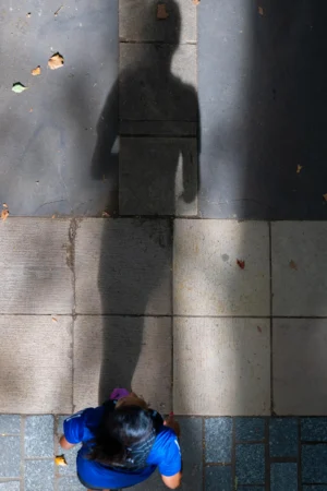 Jean-Paul · 06/09/2025 · 2000 x 3000 Cette photographie capture une silhouette d’un enfant assis sur des pavés, avec son ombre démesurément allongée projetée sur un mur. Le sujet principal est cette ombre, contrastant avec la petite taille réelle de l’enfant. La composition joue sur ce contraste saisissant entre la réalité et son reflet.Le cadrage est serré sur les pieds de l’enfant et son ombre, créant une perspective dramatique. La lumière naturelle, probablement rasante, accentue les ombres et les contrastes. Les couleurs dominantes sont les tons gris des pavés et du mur, avec des touches de bleu sur les vêtements de l’enfant.L’ambiance est à la fois poétique et mélancolique, évoquant une solitude et une grandeur inattendue.