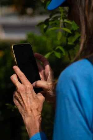 Jean-Paul · 06/09/2025 · 2000 x 3000 Cette photographie met en scène une personne âgée, sujet principal, utilisant un smartphone. Le cadrage est serré sur ses mains et son appareil, avec un arrière-plan flou de végétation.La lumière est naturelle et douce, créant des ombres légères. Les couleurs dominantes sont les tons bleus du vêtement et les verts de la végétation, avec des nuances chaudes sur les mains.Le style est réaliste et documentaire, capturant un moment du quotidien. L’ambiance est calme et intime, évoquant la connexion moderne avec la technologie à tout âge.