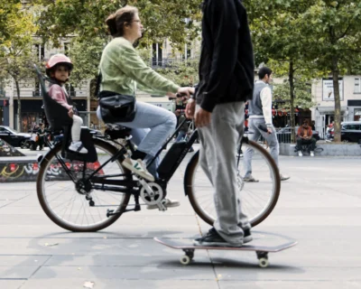 Cyrille · 13/09/2025 · 3280 x 2624 Cette photographie met en scène deux personnes en mouvement dans un espace urbain. Le sujet principal est un adulte sur un vélo avec un enfant assis devant lui, tandis qu’un autre adulte fait du skateboard à l’avant-plan.La composition est dynamique, avec un cadrage serré sur les jambes et les pieds des protagonistes, créant un effet de profondeur. La lumière semble naturelle, probablement en journée, avec des ombres douces. Les couleurs dominantes sont neutres et naturelles : gris (pavés, vêtements), vert (végétation), et touches de couleurs vives sur les vêtements et le vélo.L’ambiance est moderne et décontractée, reflétant une vie urbaine active.