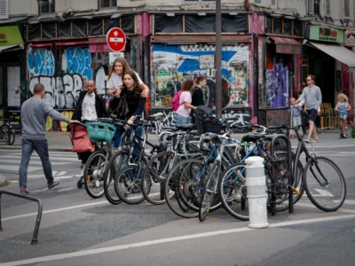 Cyrille · 13/09/2025 · 3840 x 2880 Cette photographie capture une scène urbaine animée. Le **sujet principal** est un groupe de vélos stationnés en bordure de trottoir, avec des personnes en arrière-plan.La **composition** est dynamique, avec un cadrage serré sur les vélos et une perspective légèrement basse. Les **couleurs dominantes** sont neutres (gris, noir des vélos) rehaussées par des touches vives (vêtements colorés des passants).La **lumière** semble naturelle, probablement diurne, avec des ombres douces. L’ambiance est **vivante et quotidienne**, reflétant une ville active et multiculturelle. Le style est **documentaire**, réaliste et sans artifice.