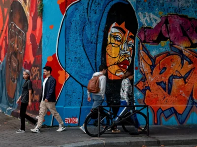 Cyrille · 13/09/2025 · 3713 x 2785 Cette photographie met en scène un cycliste au premier plan, vêtu d’une veste sombre, passant devant un mur coloré. Le sujet principal est ce mur couvert de graffitis aux couleurs vives et contrastées, dominées par des tons bleus, rouges et oranges, avec un visage stylisé en relief.La composition joue sur la profondeur avec les deux piétons en arrière-plan. Le cadrage est serré, mettant en valeur les détails du graffiti et l’action du cycliste. La lumière semble naturelle, créant des ombres douces. L’ambiance est urbaine, dynamique et artistique, mêlant mouvement et créativité.