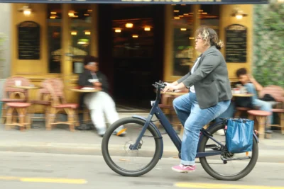 Francoise · 13/09/2025 · 3840 x 2560 Cette photographie met en scène une femme comme sujet principal, assise sur un vélo électrique en ville. La composition montre un premier plan dynamique avec elle, tandis qu’en arrière-plan, on aperçoit un café animé avec des tables et des clients.Le cadrage est serré sur la cycliste, avec une légère flou artistique pour l’arrière-plan. La lumière semble naturelle, probablement en journée, avec des ombres douces. Les couleurs dominantes sont les tons neutres (gris, bleu) et quelques éclats de couleurs vives comme les chaussures roses de la femme.L’ambiance est urbaine et détendue, évoquant une pause ou un trajet quotidien dans une ville animée.