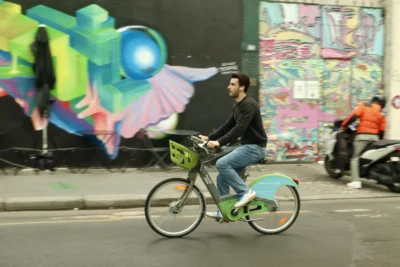 Francoise · 13/09/2025 · 3840 x 2560 Cette photographie montre un homme comme sujet principal, pédalant un vélo électrique coloré (bleu et vert) dans une rue urbaine. La composition met en valeur le cycliste au premier plan, avec un cadrage serré. En arrière-plan, un mur couvert de graffitis colorés et lumineux attire l’attention.La lumière semble naturelle, probablement prise en soirée ou de nuit, avec des éclairages artificiels. Les couleurs dominantes sont vives : bleu électrique, vert fluo, et teintes vives des graffitis. Le style est urbain et dynamique, créant une ambiance jeune et énergique, mêlant mouvement et art de rue.