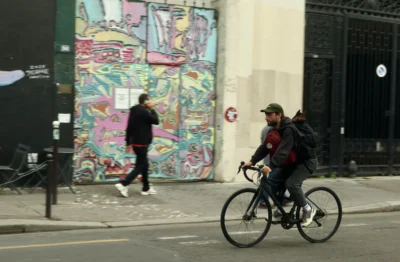 Francoise · 13/09/2025 · 3840 x 2516 Cette photographie montre un cycliste comme sujet principal, vêtu d’un manteau sombre et d’un sac à dos, en mouvement. La composition inclut aussi deux personnes en arrière-plan près d’un mur couvert de graffitis colorés et variés.Le cadrage est serré sur le cycliste, avec une profondeur de champ qui met en valeur les détails du premier plan et les éléments urbains en arrière-plan. La lumière semble naturelle, un peu grise, typique d’un jour couvert. Les couleurs dominantes sont les teintes vives des graffitis contrastant avec les tons neutres de la ville.L’ambiance est urbaine et dynamique, mêlant art de rue et vie quotidienne.