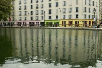 Francoise · 13/09/2025 · 3840 x 2560 Cette photographie met en scène un bâtiment urbain reflété dans une étendue d’eau calme. Le sujet principal est la symétrie entre le reflet des façades et leur image inversée.La composition repose sur une ligne horizontale nette séparant l’eau et les bâtiments. Le cadrage est serré, centré sur les fenêtres et les enseignes. La lumière est douce, probablement naturelle, créant des contrastes subtils. Les couleurs dominantes sont les tons neutres (beige, gris) avec des touches de vert et de jaune.Le style est épuré et photographique, évoquant une ambiance paisible et presque onirique, typique des scènes urbaines apaisées.