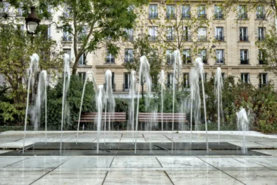 Frédéric · 13/09/2025 · 2518 x 1680 Cette photographie met en scène un **jeu de fontaines urbaines** comme sujet principal. La composition est symétrique, avec des jets d’eau alignés et un bassin central.Le cadrage est serré sur les fontaines, mais inclut en arrière-plan des bâtiments et des arbres. La lumière est naturelle, douce et diffuse, probablement en journée. Les couleurs dominantes sont des **tons froids** (bleu-gris de l’eau, gris des structures) contrastant avec le **vert des arbres** et les **teintes chaudes des façades**.Le style est épuré et contemporain, évoquant une ambiance **calme et élégante**, typique d’un espace public urbain bien entretenu.