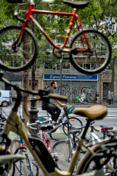 Frédéric · 13/09/2025 · 1120 x 1680 Cette photographie met en scène un cycliste en mouvement, flou, au premier plan. La composition montre une scène urbaine avec des vélos stationnés en avant et un fond de rue animée. Le cadrage est serré, avec une perspective légèrement basse, accentuant la dynamique du sujet.Les couleurs dominantes sont les tons froids (gris, bleu) et chauds (rouge du vélo, vert des arbres). La lumière semble naturelle, avec des contrastes modérés. Le style est documentaire et spontané, capturant un instant de vie quotidienne. L’ambiance est vivante et urbaine, mêlant mouvement et immobilité.